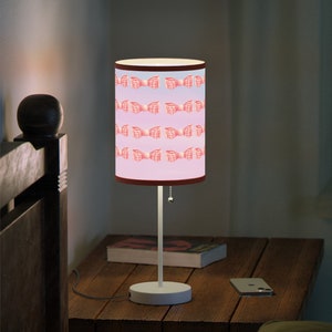 May include: A white table lamp with a pink and white patterned lampshade featuring a repeating bow design. The lamp is on a wooden nightstand with a book and a cell phone.