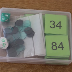 Könnte beinhalten: Eine transparente Plastikbox mit grünen und weißen Würfeln und grünen Zahlkarten. Die Karten zeigen die Zahlen 34 und 84.