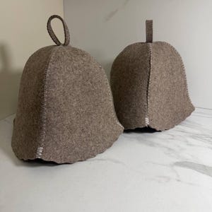 May include: Two taupe-colored sauna hats. The hats are bell-shaped with a rounded top and a small loop or tab for hanging. The hats are made of a felt-like material and have white stitching along the seams.