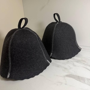 May include: Two dark gray sauna hats with a bell shape and a loop at the top. The hats have white stitching along the seams and edges. The hats are made of a felt-like material.