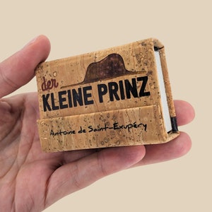May include: A small, cork-covered box with the title "der KLEINE PRINZ" and the author's name "Antoine de Saint-Exupéry" printed on the front.