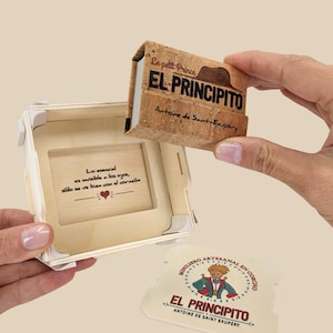 Puede incluir: Una caja de madera con un libro de corcho en su interior. El libro se titula "El Principito" de Antoine de Saint-Exupéry. La caja tiene una cita del libro: "Lo esencial es invisible a los ojos, solo se ve bien con el corazón".