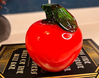 May include: A red glass apple with a green glass leaf on top. The apple is sitting on a black book with gold lettering.