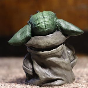 May include: A small green and gray figurine of a character from the Star Wars franchise. The figurine is wearing a brown robe and has large ears.