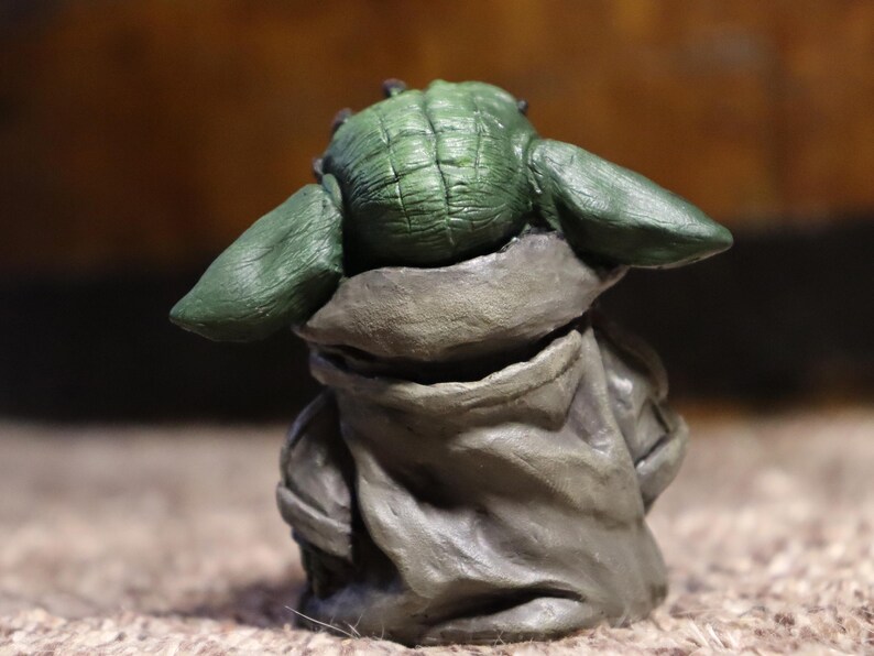 May include: A small green and gray figurine of a character from the Star Wars franchise. The figurine is wearing a brown robe and has large ears.