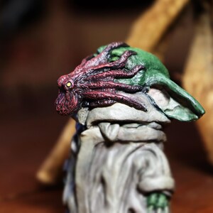 May include: A small, green and gray figurine with a red and purple octopus on its head. The figurine is made of a textured material and has a detailed design.