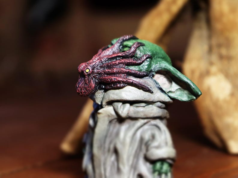 May include: A small, green and gray figurine with a red and purple octopus on its head. The figurine is made of a textured material and has a detailed design.