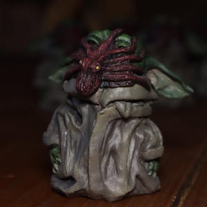 May include: A small green figurine with a red octopus on its head. The figurine is wearing a grey robe and has a green face.
