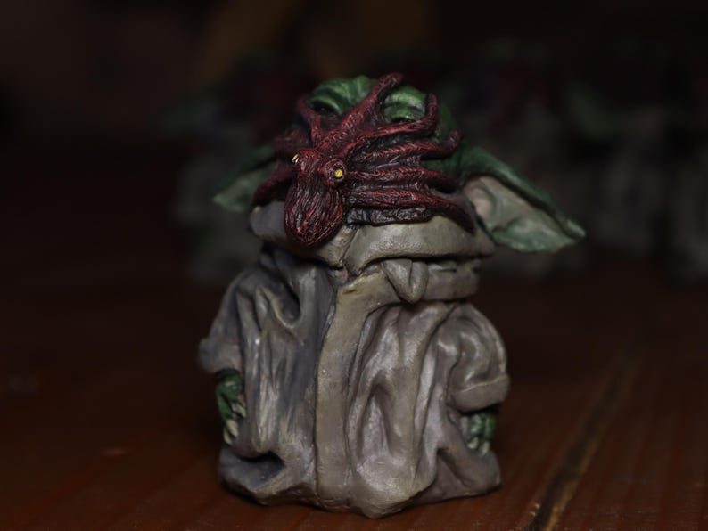 May include: A small green figurine with a red octopus on its head. The figurine is wearing a grey robe and has a green face.