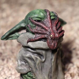 May include: A green and gray figurine of a character wearing a red and brown octopus mask.