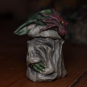 May include: A small, green and grey figurine with a red and purple tentacle-like head. The figurine is standing on a wooden surface.