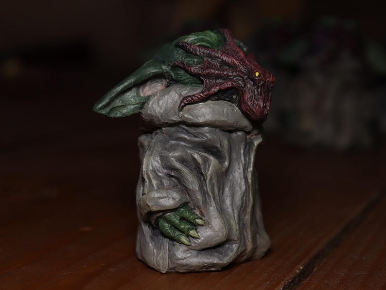 May include: A small, green and grey figurine with a red and purple tentacle-like head. The figurine is standing on a wooden surface.