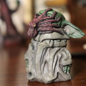 May include: A small, green and grey figurine of a character wearing a red and brown octopus on its head. The figurine is sitting on a wooden surface.