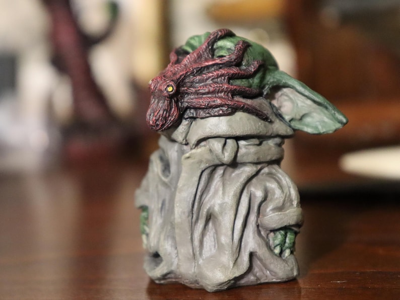 May include: A small, green and grey figurine of a character wearing a red and brown octopus on its head. The figurine is sitting on a wooden surface.