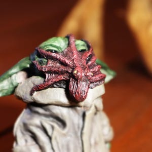 May include: A small green and gray figurine with a red octopus on its head. The octopus has a textured surface and is a vibrant red color.