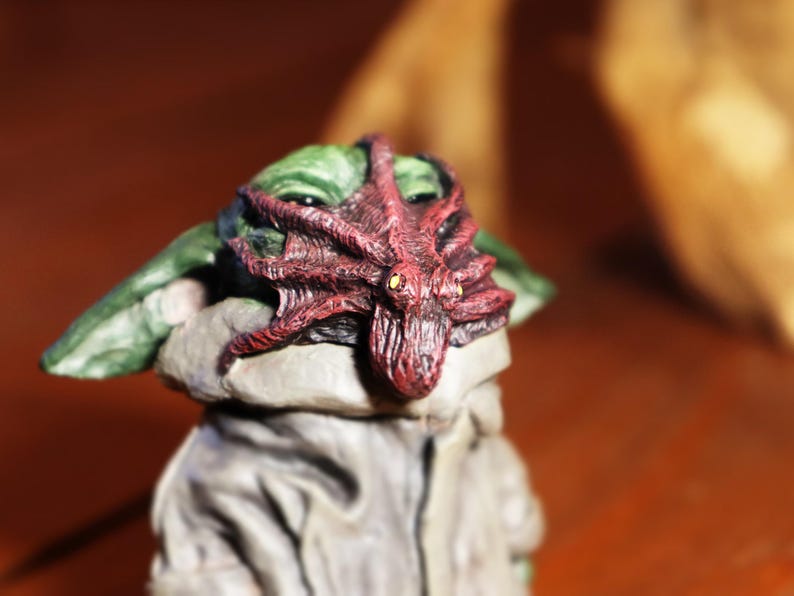May include: A small green and gray figurine with a red octopus on its head. The octopus has a textured surface and is a vibrant red color.