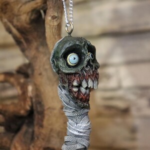 Può includere: Una collana con catena d'argento con un ciondolo a forma di testa di zombie verde e rossa con un occhio bianco. La testa di zombie è avvolta in bende bianche.