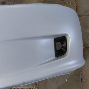 May include: A light gray car bumper with a black fog light housing. The bumper has a smooth, curved design and a small indentation. The fog light housing is rectangular with a rounded opening.