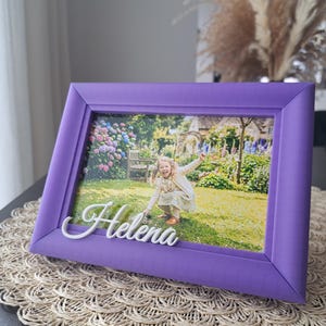 Könnte beinhalten: Ein lila Bilderrahmen mit einem Foto eines kleinen Mädchens in einem Garten. Der Name "Helena" ist in weißer Schrift auf dem Rahmen geschrieben. Der Rahmen liegt auf einem gewebten Platzset.