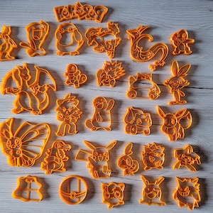 Könnte beinhalten: Ein Set mit 21 orangefarbenen Plastik-Ausstechern mit verschiedenen Pokémon-Charakteren, darunter Pikachu, Bisasam, Glumanda, Schiggy und viele mehr. Die Ausstecher sind in einem Raster auf einer hellbraunen Holzoberfläche angeordnet.