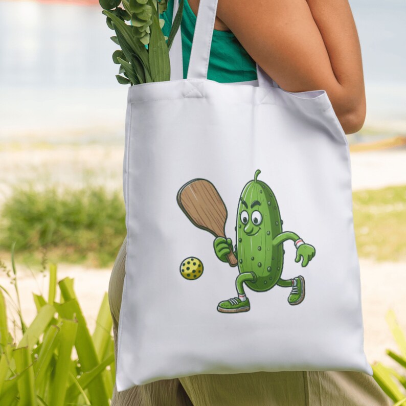 May include: White tote bag with a cartoon pickle holding a paddle and a yellow ball. The pickle is wearing sneakers and has a green body with a brown face.