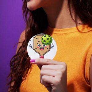 May include: A white sticker with a cartoon illustration of a wooden paddleball racket with a smiling face and a green paddleball. The paddleball is above the racket and appears to be bouncing off of it.