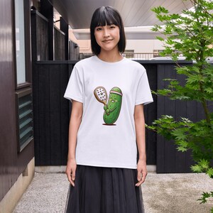 May include: A white t-shirt with a cartoon illustration of a green pickle holding a tennis racket. The pickle is wearing a pink headband and has a smiling face.