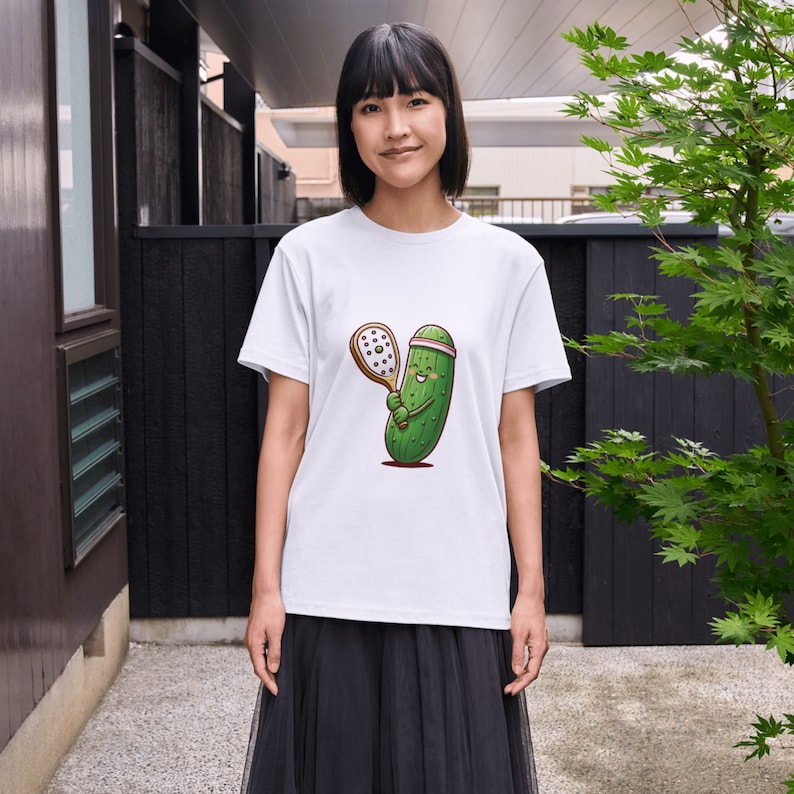 May include: A white t-shirt with a cartoon illustration of a green pickle holding a tennis racket. The pickle is wearing a pink headband and has a smiling face.