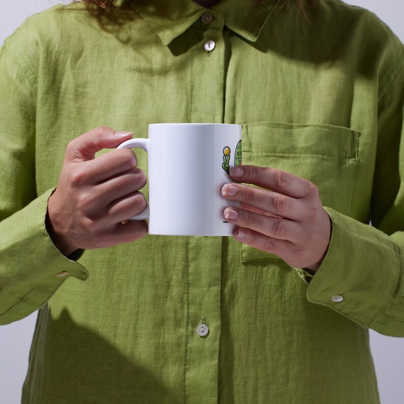 May include: A white ceramic mug with a green cactus illustration and the text "Cactus Queen". The mug is being held by a person wearing a green shirt.