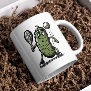 May include: White ceramic mug with a cartoon illustration of a green pickle playing tennis. The pickle is holding a tennis racket and has a happy expression on its face. The mug has a black handle.
