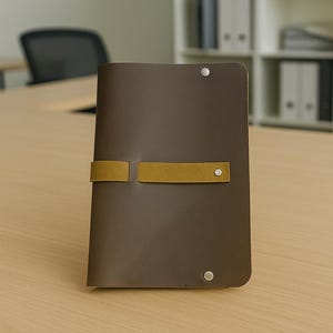 以下が含まれることがあります： マスタード色のストラップ留めが付いた茶色の革製ノート。 ノートは閉じられ、ストラップと金属リベットで固定されています。 ノートは明るい木製の表面にあります。