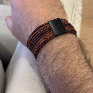 K&ouml;nnte beinhalten: Geflochtenes Lederarmband in Braun mit schwarzem Magnetverschluss. Das Armband besteht aus mehreren Lederstr&auml;ngen in verschiedenen Braunt&ouml;nen. Das Armband ist um ein Handgelenk gewickelt.
