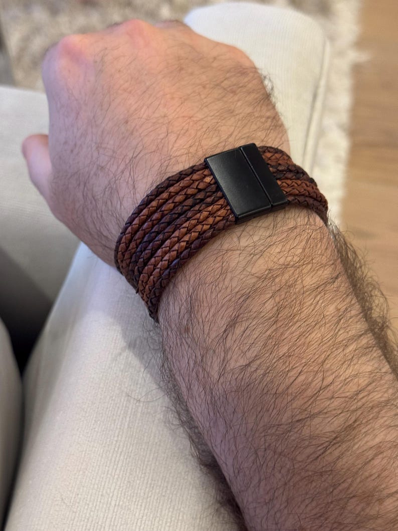 K&ouml;nnte beinhalten: Geflochtenes Lederarmband in Braun mit schwarzem Magnetverschluss. Das Armband besteht aus mehreren Lederstr&auml;ngen in verschiedenen Braunt&ouml;nen. Das Armband ist um ein Handgelenk gewickelt.