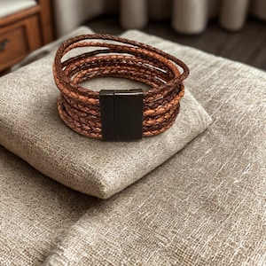 K&ouml;nnte beinhalten: Ein braunes Lederarmband mit mehreren geflochtenen Str&auml;ngen. Das Armband hat einen Magnetverschluss und liegt auf einem beigen Kissen. Die Lederstr&auml;nge sind in hellen und dunklen Braunt&ouml;nen gehalten.
