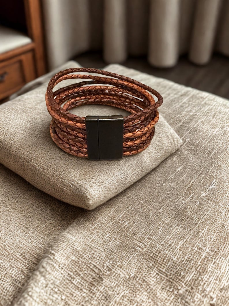 K&ouml;nnte beinhalten: Ein braunes Lederarmband mit mehreren geflochtenen Str&auml;ngen. Das Armband hat einen Magnetverschluss und liegt auf einem beigen Kissen. Die Lederstr&auml;nge sind in hellen und dunklen Braunt&ouml;nen gehalten.