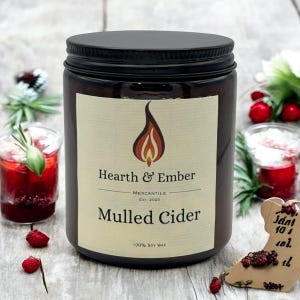 May include: A brown glass candle jar with a black lid and a white label. The label says "Hearth & Ember Mercantile Est. 2023 Mulled Cider 100% Soy Wax".