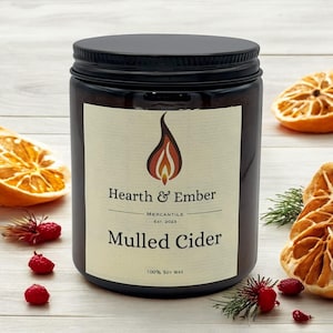 May include: A brown glass candle jar with a black lid and a label that reads "Hearth & Ember Mercantile Est. 2023 Mulled Cider 100% Soy Wax". The candle is surrounded by dried orange slices and sprigs of greenery.