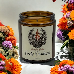 Könnte beinhalten: Ein braunes Glasgefäß mit einer Kerze und einem weißen Etikett. Das Etikett zeigt eine Schwarzweißillustration einer Flamme mit einem Kranz darum. Der Text auf dem Etikett lautet "Hearth & Ember Lady Danbury 100% Soy Wax".
