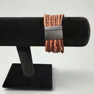 K&ouml;nnte beinhalten: Ein mehrstr&auml;ngiges Lederarmband in Braun- und Beiget&ouml;nen, mit einem rechteckigen, dunklen Metallverschluss. Das Armband wird auf einem schwarzen Samt-Schmuckst&auml;nder pr&auml;sentiert, der seine Textur und Farbvariationen hervorhebt.