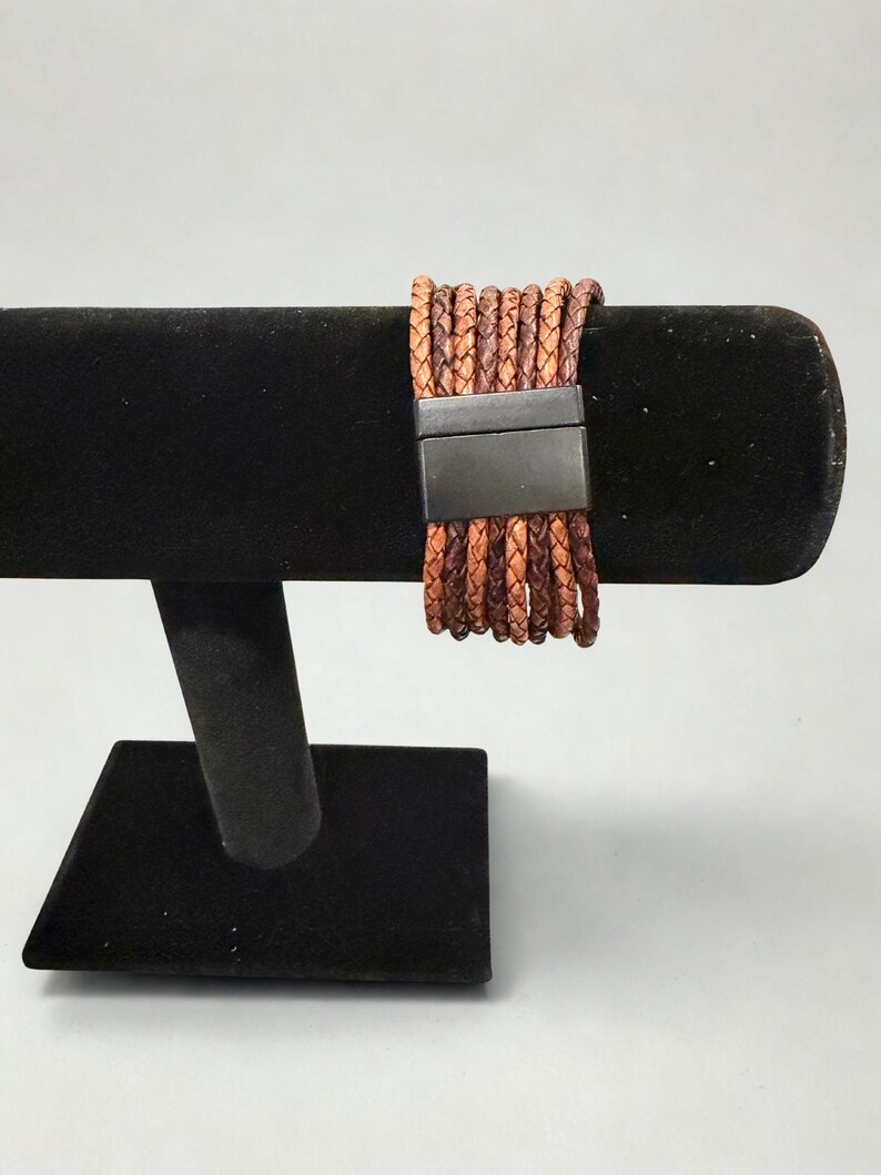 K&ouml;nnte beinhalten: Ein mehrstr&auml;ngiges Lederarmband in Braun- und Beiget&ouml;nen, mit einem rechteckigen, dunklen Metallverschluss. Das Armband wird auf einem schwarzen Samt-Schmuckst&auml;nder pr&auml;sentiert, der seine Textur und Farbvariationen hervorhebt.