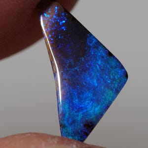 May include: A polished, triangular opal gemstone with a vibrant display of blues, greens, and hints of gold. The stone's surface reflects light, showcasing a dynamic play of color and a unique pattern. The opal is held between fingers.