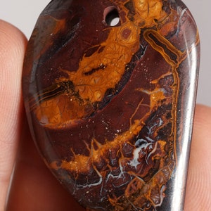 May include: A teardrop-shaped pendant with a drilled hole at the top. The stone features a mix of dark brown, orange, and white patterns. The polished surface highlights the natural veining and color variations of the stone.
