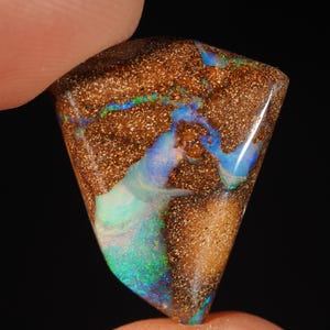 Könnte beinhalten: Ein dreieckiger Boulder-Opal-Edelstein mit einer texturierten, erdbraunen Matrix und lebendigen, irisierenden Adern in Blau, Grün und Violett, die durch die Mitte und entlang der Kanten der polierten Steinoberfläche verlaufen.