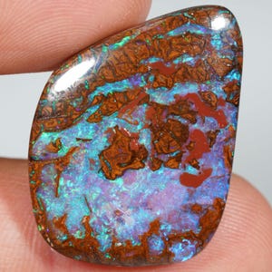 Puede incluir: Una piedra preciosa pulida con un patrón único de tonos marrones, azules, rojos y morados. La superficie de la piedra tiene un aspecto texturizado, con una mezcla de colores que crea una exhibición llamativa. La piedra preciosa se sostiene entre los dedos.