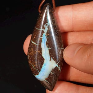 May include: A teardrop-shaped pendant with a brown base color and a striking pattern of blue and white opal. The opal has a unique, crackled appearance with thin, light blue veins. The pendant is suspended from a brown leather cord.