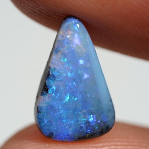 May include: A teardrop-shaped opal gemstone with a vibrant display of blues and greens. The polished surface reflects light, showcasing internal patterns and color variations. The stone is held between fingers, highlighting its size and shape.