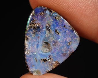 May include: A triangular-shaped opal gemstone with a blue and white color scheme, speckled with black and gold flecks. The stone has a unique pattern and a hole in the center. The opal is held between two fingers against a black background.