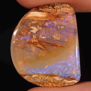 Pode incluir: Uma pedra preciosa de opala boulder polida com uma forma suave e arredondada. A pedra apresenta uma base translúcida de cor de mel com flashes vibrantes de luz azul e roxa iridescente, intercalados com inclusões naturais de matriz de ferro.