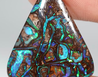 May include: A triangular-shaped gemstone with a unique pattern of brown, blue, and green hues. The stone's surface displays a mosaic-like arrangement of colors, creating a striking visual effect. The gemstone is held by a finger.