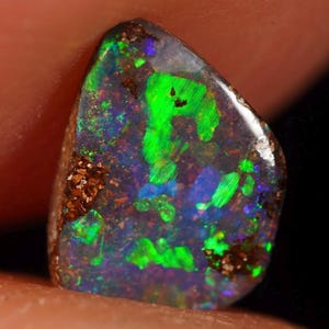 Op de afbeelding: Een gepolijste opaal edelsteen met een donkere basiskleur, met levendige flitsen van groen, blauw en paars. De steen heeft een ruw, getextureerd oppervlak met bruine insluitsels en is onregelmatig van vorm.